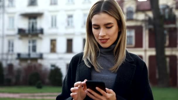 Attractive woman typing Messages on her Smartphone while having a standing in the Street. Having fun and enjoying time outdoor. Pretty brunette with Smartphone in front of Camera. Beautiful girl - Materiał filmowy, wideo