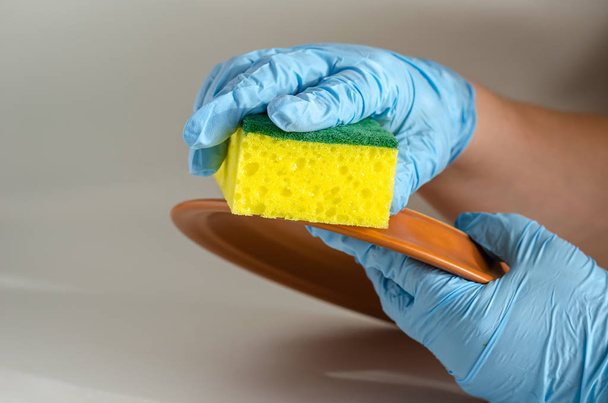 Τα χέρια με μπλε προστατευτικά γάντια πλένουν το πιάτο. Ένα πλυντήριο πιάτων δουλεύει στην κουζίνα με βρώμικα πιάτα. Πυροβολισμός στο ύψος των ματιών. Κοντινό πλάνο. Επιλεκτική εστίαση. Διάταξη φωτογραφίας τοπίου. - Φωτογραφία, εικόνα