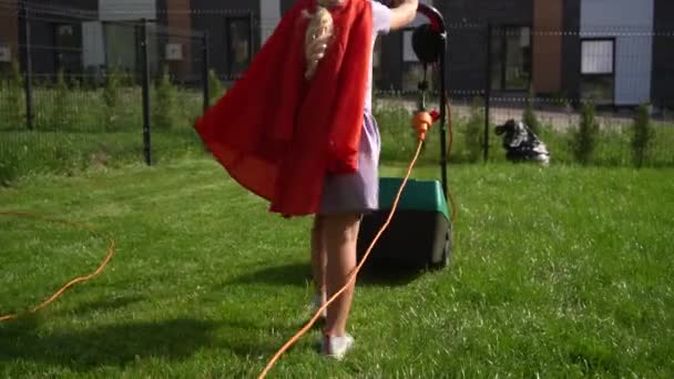 Super girl girl in red cloak mowing lawn in fenced house yard. Corded lawn mower - Footage, Video