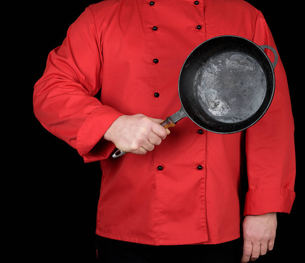 cook in red uniform holding an empty round black frying pan, black background - Foto, Imagen