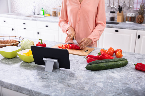 Nő használ digitális tabletta vágás közben Red Bell Pepper vágódeszka a konyhában - Fotó, kép