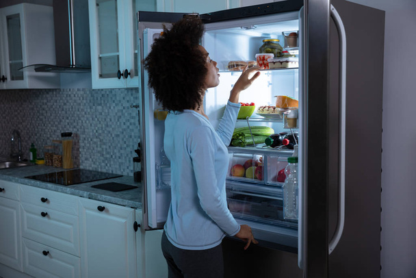 Side View Of Young African Woman Searching Food In An Open Refrigerator - Photo, image
