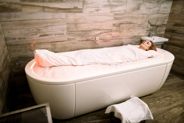 Side angle view of pretty young woman lying in the hot spa capsule, wrapped with special cover during the treatment procedure in modern spa medical center - Photo, Image