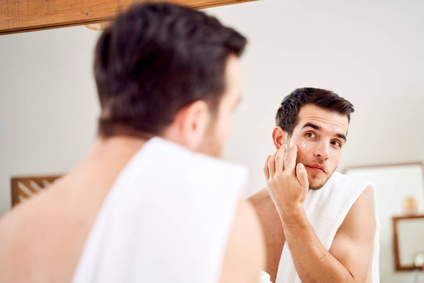 Male spreads his face with white cream while standing in front of mirror in bath - Foto, Imagen