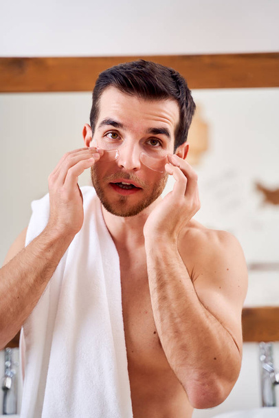 Brunet male applies transparent patches under his eyes while standing in bath opposite mirror - Foto, Bild
