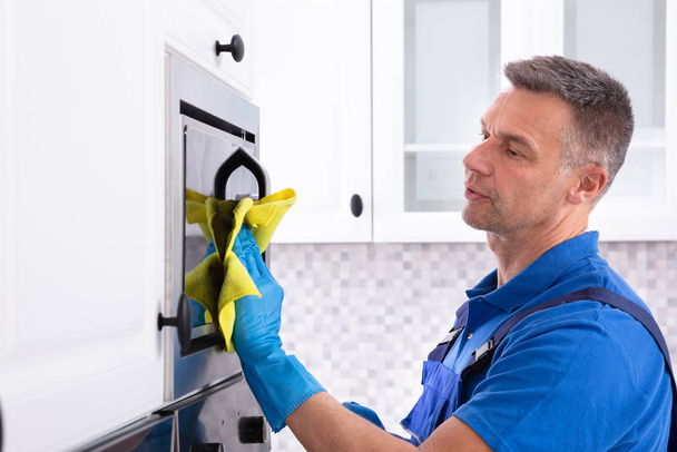 Side View Of Male Janitor Cleaning Oven With Yellow Napkin In Kitchen - Foto, imagen