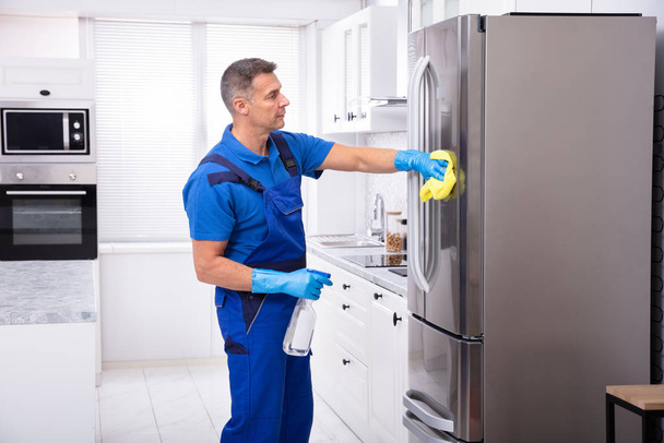 Male Janitor Cleaning Refrigerator With Yellow Napkin And Spray Detergent - Фото, изображение
