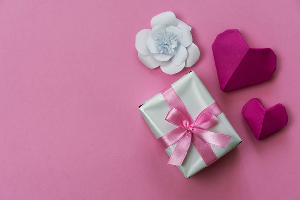 Valentine's Day background with a gift box wrapped with pink ribbon, white paper flowers and origami hearts, copy space on left side. - Фото, изображение