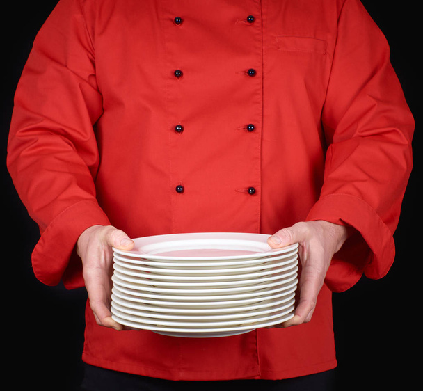 cook in red uniform holds in his hands a stack of round white empty plates, black background - Fotó, kép