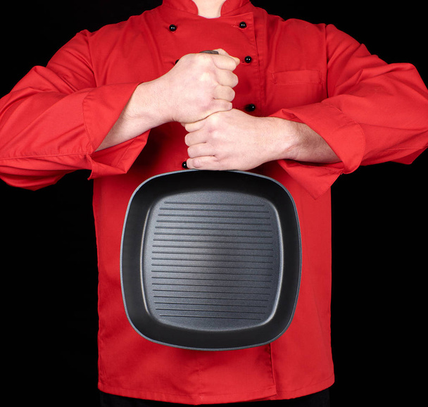 cook in red uniform holding an empty square black frying pan, black background - Fotó, kép