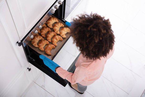 A Happy Young Woman Wearing Blue Mitt Removing Baked Croissants Tray From An Oven - Фото, зображення