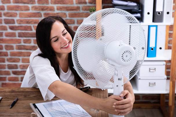 Smiling Young Woman Enjoying Breeze With Electric Fan At Office - Photo, Image