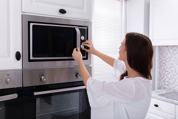 Side View Of A Young Woman Using Microwave Oven In Kitchen - Foto, immagini