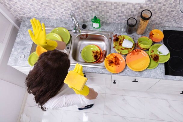 Sad Young Woman Looking At Dirty Utensils Near Sink In Kitchen - Фото, изображение