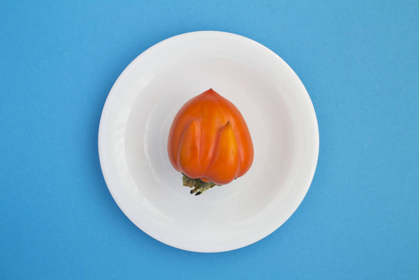 Ugly food. Ugly persimmon on the white plate  in the center of the  blue background. Top view. Copy space. - Photo, Image