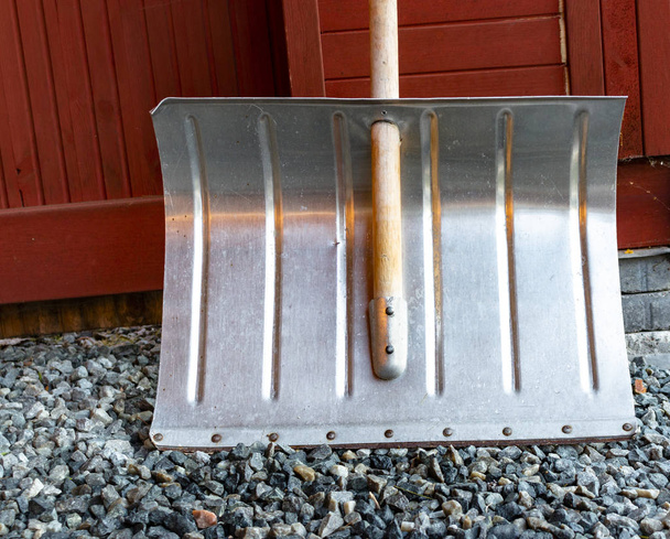 Silver-colored metal snow shovel leans against a red wooden wall - 写真・画像