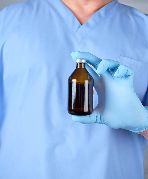 doctor in blue uniform and latex gloves holding a brown glass bottle with medicine for treatment - Photo, image