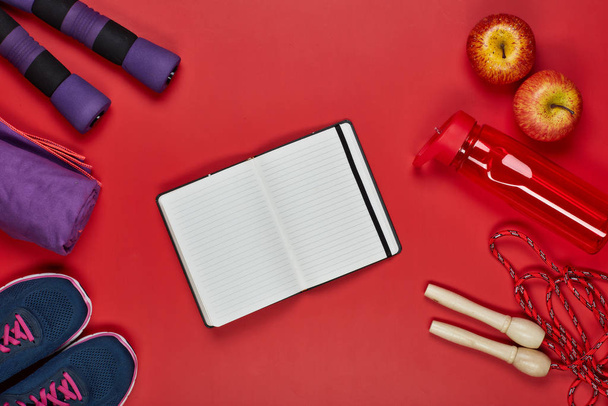 Female fitness still life. Sport accessories: dumbbells, rope, sneakers and blank sheet in notebook on a red background. Top view. Fitness, sport and healthy lifestyle concept. - 写真・画像