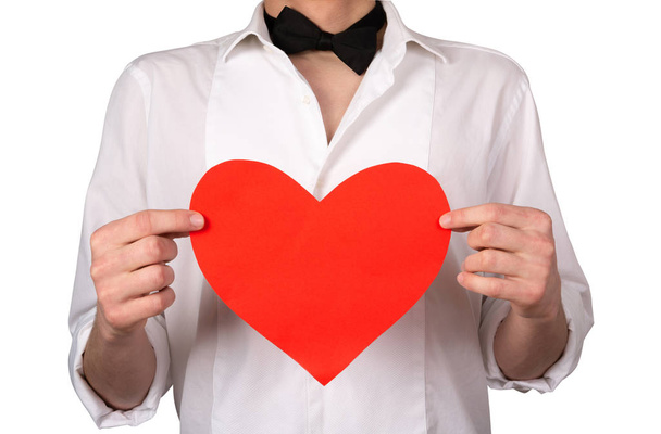 A man in a white shirt with a bow tie holds a red heart. Closeup of a young man without a face holding an empty paper heart, on a white background. Front view, place for text, copy space, isolate. - Fotoğraf, Görsel