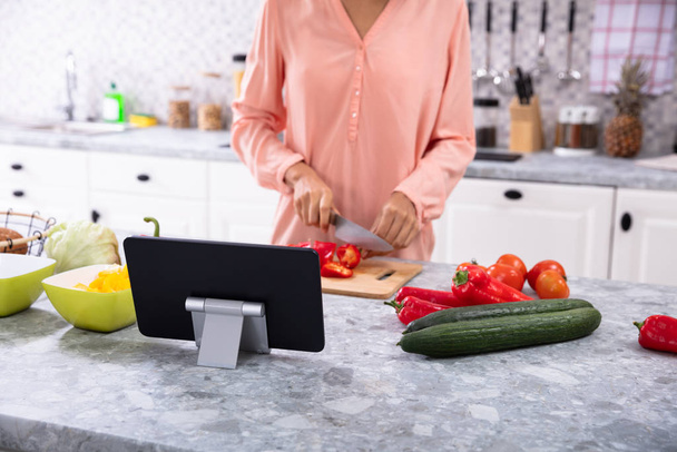 Nainen, joka käyttää digitaalista tablettia leikkaamalla Red Bell Pepper on leikkuulauta keittiössä
 - Valokuva, kuva