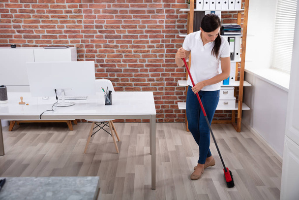 Happy Young Female Janitor Cleaning Floor With Broom In Office - 写真・画像