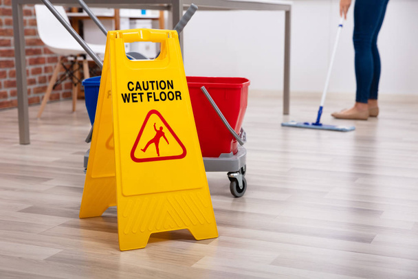 Low Section Of Female Cleaner Standing In Office With Mop And Caution Wet Floor Sign - Φωτογραφία, εικόνα