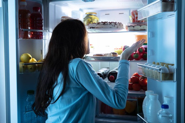 Rear View Of A Young Woman Standing In Front Of An Open Refrigerator Shrugging - Photo, Image