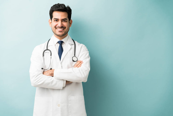 Smiling Hispanic doctor standing with arms crossed against blue background - Foto, imagen