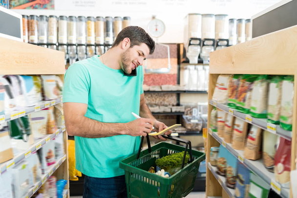 Sonriente cliente masculino que comprueba la lista de compras mientras habla en el teléfono inteligente en el supermercado
 - Foto, Imagen