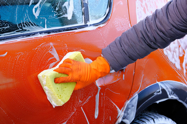 Czyszczenie piwami mydlanymi w samoobsługowej myjni samochodowej. Mężczyzna w gumowych rękawiczkach myje pomarańczowy samochód żółtą gąbką. mężczyzna ręka i samochód ciało zbliżenie. - Zdjęcie, obraz