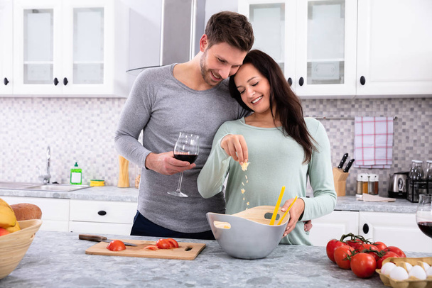 Smiling Wife Sprinkling Butter With Her Husband Standing In Kitchen Holding Wine Glass - 写真・画像