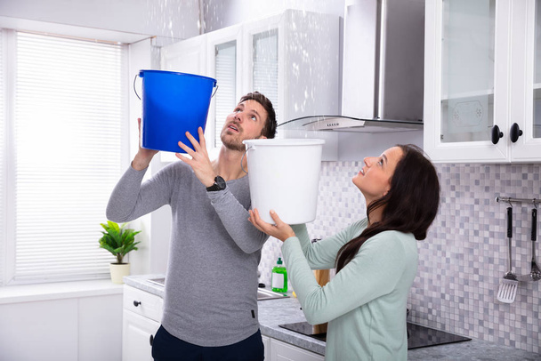 Close-up Of A Worried Young Couple Collecting Water Leaking From Ceiling In The Blue Bucket - Foto, Imagem