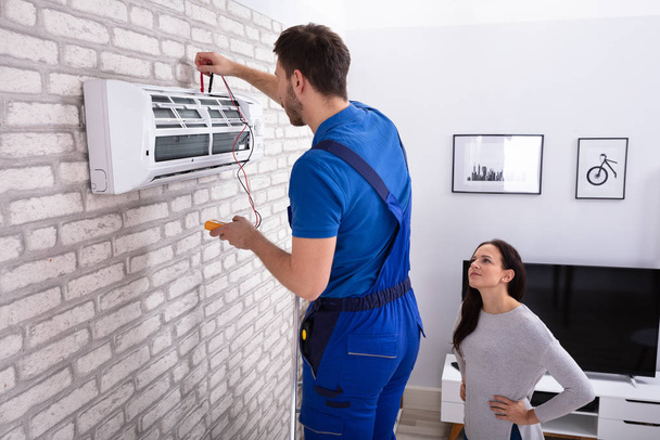 Smiling Woman Looking At Male Technician Repairing Air Conditioner With Digital Multimeter At Home - Photo, Image