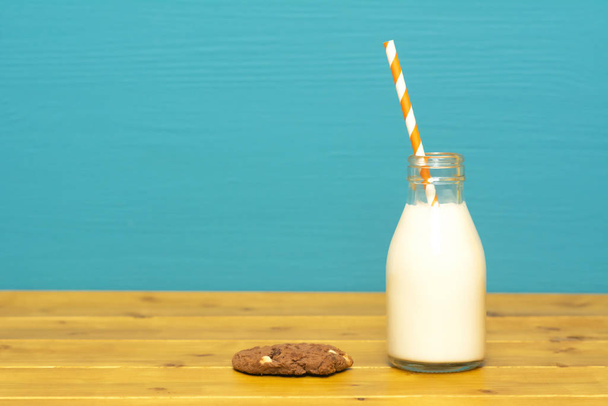 Fresh creamy milk with a retro paper straw in a one-third pint glass milk bottle and a chocolate chip cookie, on a wooden table against a teal background - Foto, imagen