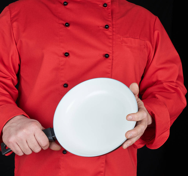 cook in red uniform holding an empty round white frying pan, black background - Photo, Image