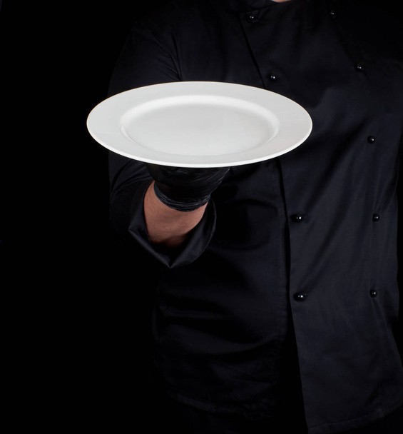 cook in black uniform  holds in his hand a round empty white plate - Fotoğraf, Görsel