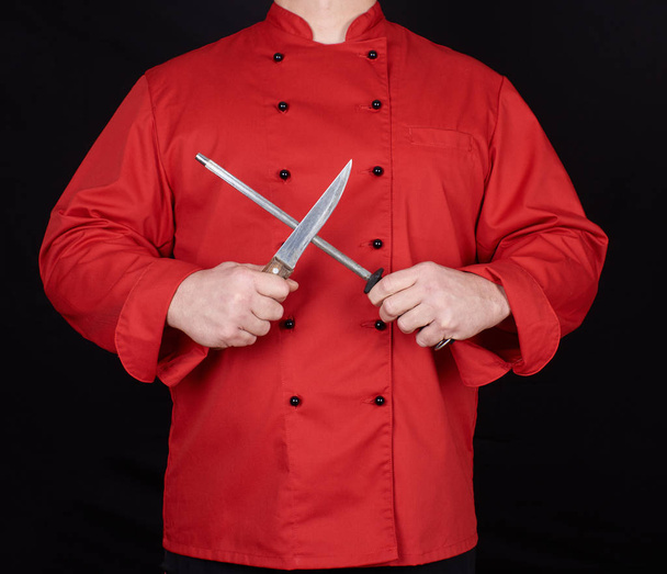 chef in red uniform sharpens a knife, black background - 写真・画像