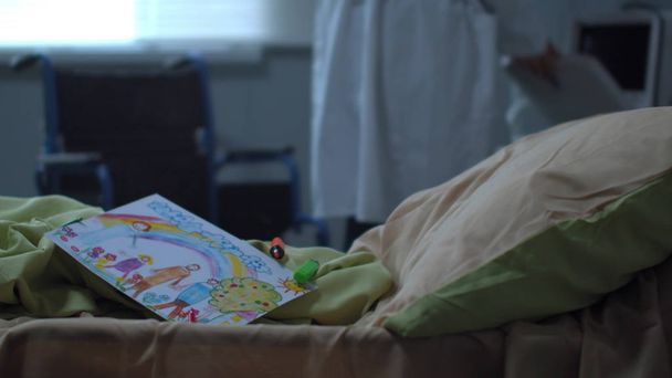 Enfoque en la cama con dibujo del niño, el médico camina sobre el fondo
 - Foto, Imagen