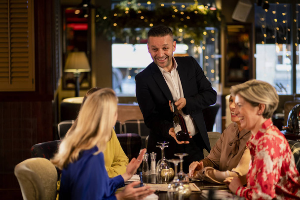 Mature waiter reccomending a bottle of champagne to a small group of mature women. They are sitting at a table in a restaurant. - Foto, Imagen