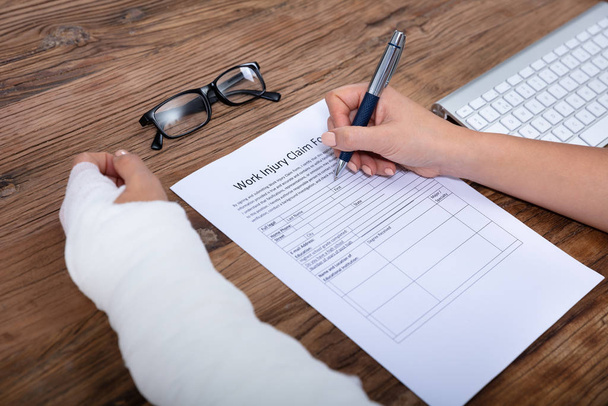 Close-up Of Businesswoman With White Bandage Hand Filling Work Injury Claim Form On Wooden Desk - 写真・画像