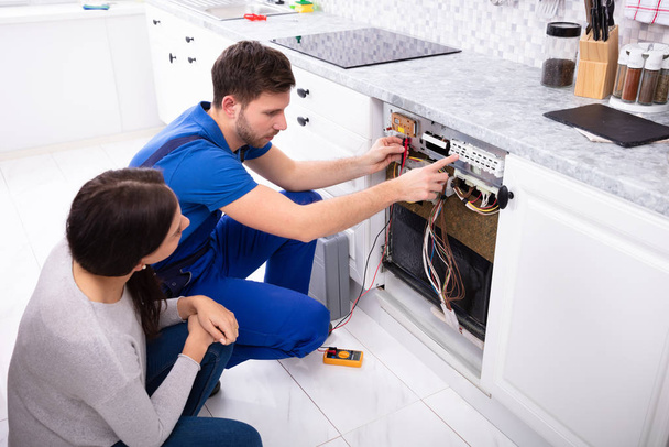 Woman Looking At Male Technician Checking Dishwasher With Digital Multimeter In Kitchen - Фото, изображение