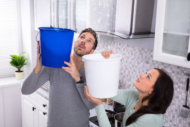 Close-up Of A Worried Young Couple Collecting Water Leaking From Ceiling In The Blue Bucket - Foto, Imagen