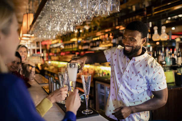 Bartender filling up a mature womans glass with prosecco. She is sitting at the bar counter with her friends. - Photo, image