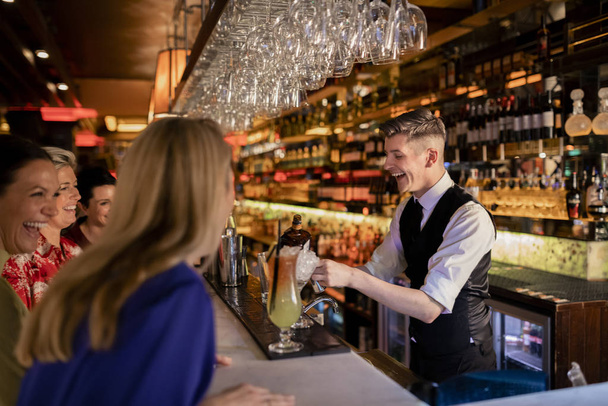 Mladý barman připravuje koktejl pro malou skupinku žen, které si užívají dámskou jízdu. Všichni se smějí a vedou konverzaci. - Fotografie, Obrázek