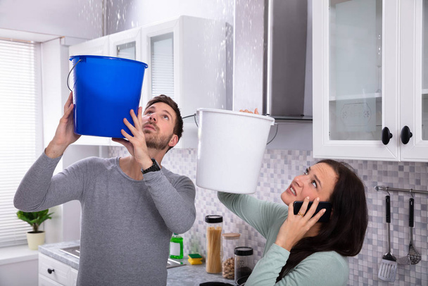 Worried Young Woman Calling Plumber On Cellphone While Man Collecting Leakage Water From Ceiling - Foto, immagini