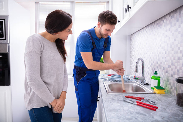 Happy Woman Looking At Male Plumber Using Plunger In The Kitchen Sink - Foto, imagen