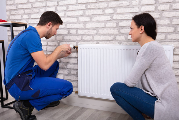 Beautiful Woman Looking At Young Male Plumber In Uniform Installing Radiator With Screwdriver - Foto, imagen