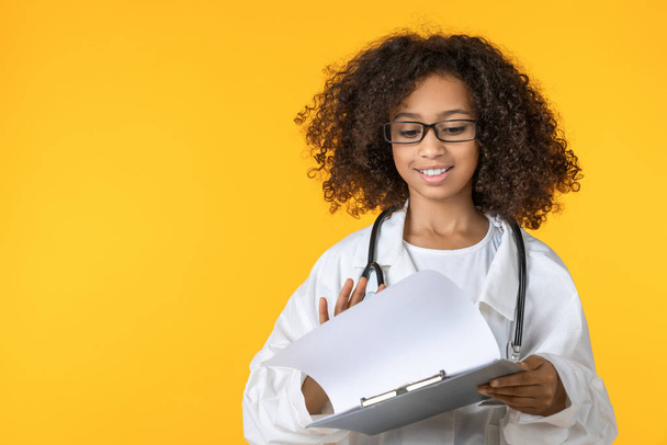 Cute little girl dressed like doctor checking notepad isolated on yellow background - Foto, Imagen