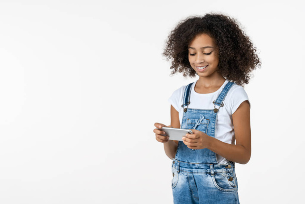 Happy little african girl playing on mobile phone standing isolated over white background - Φωτογραφία, εικόνα