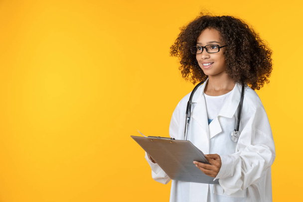 Cute little girl dressed like doctor with notepad isolated on yellow background - Foto, Imagen